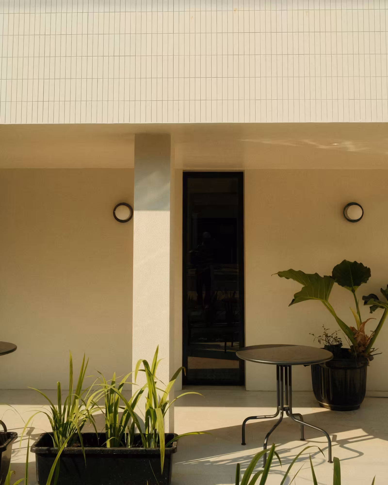 Sonnige Terrasse mit Rundtisch, Pflanzen und schwarzer Tür an heller Hauswand